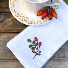 Handgefertigte Herbstserviette mit Hagebutte aus weißem Leinen von Atelier Luhabru, naturgetreu bestickt, mit Kaffeetasse und echten Hagebutten auf Holztisch